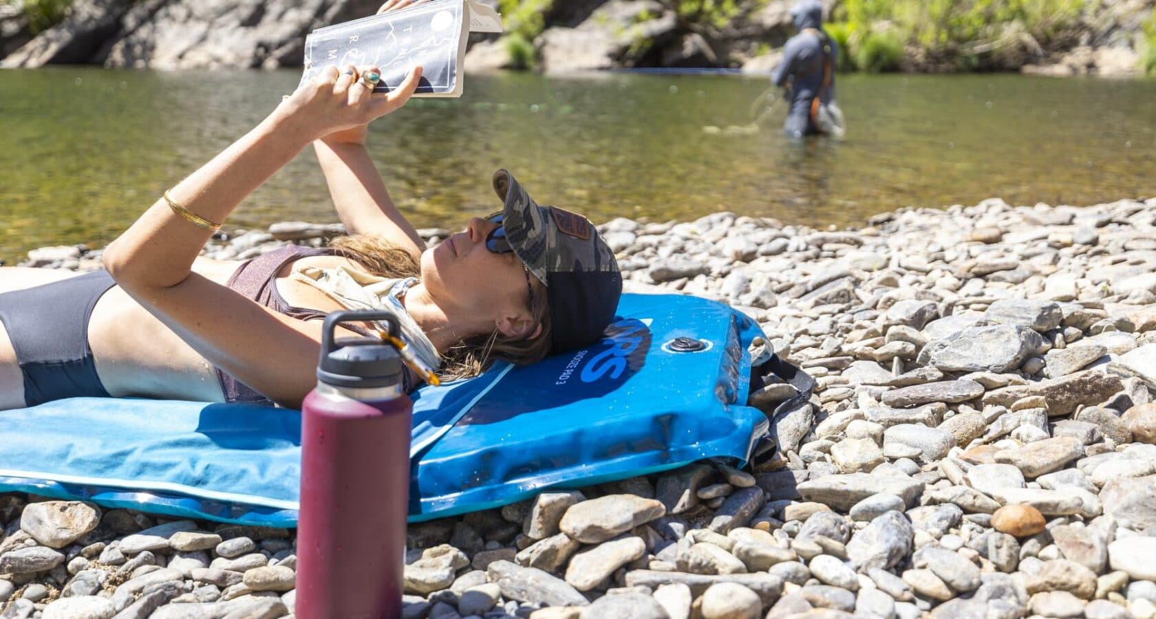 A woman reading a book and lying on her back on an inflatable blue pad situated on a rocky shore next to a river where a man is fishing in the background.