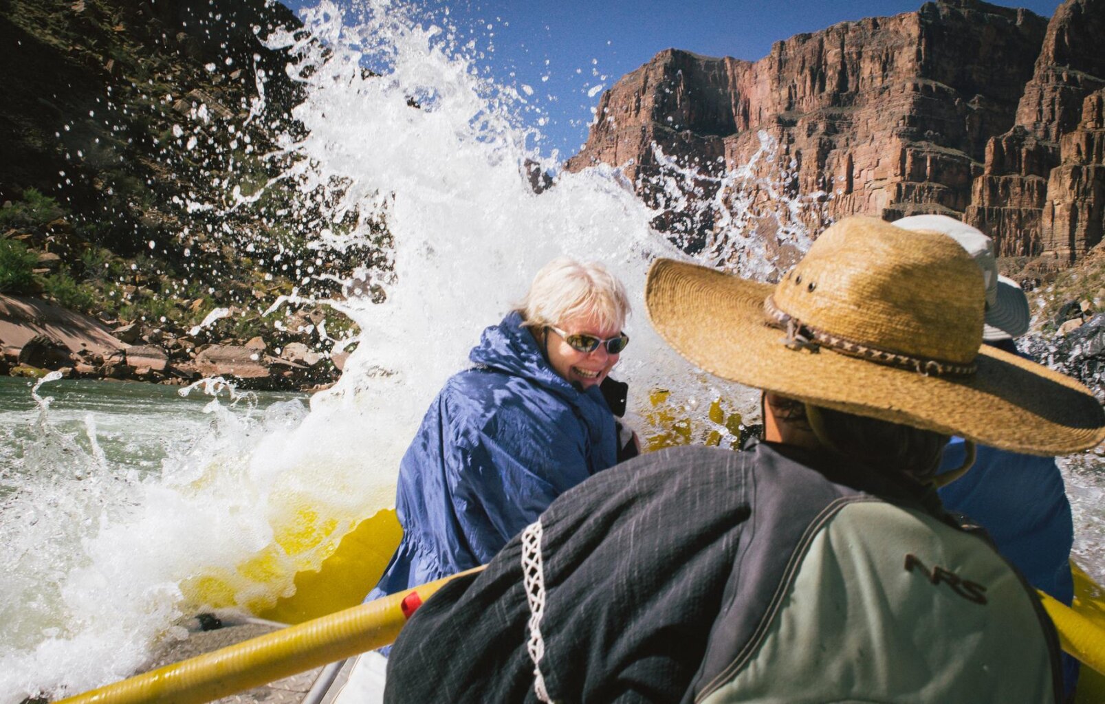 A woman turns away from a large splash coming over the front of the yellow raft she's riding on during a rafting trip through Grand Canyon