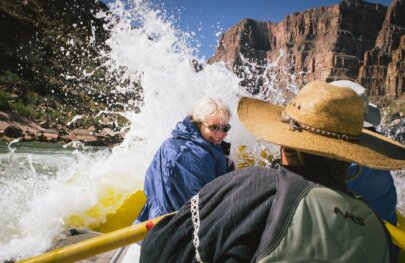 A woman turns away from a large splash coming over the front of the yellow raft she's riding on during a rafting trip through Grand Canyon