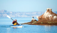 A woman kayaks on the Sea of Cortez in Baja Mexico and looks at a seagull standing on a nearby rock formation.