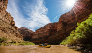 A yellow raft with people floats down the San Juan River with a sunburst above the canyon walls