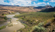 A vast stretch of land with mountains in the background and a yellow raft rowing down the Owyhee River.