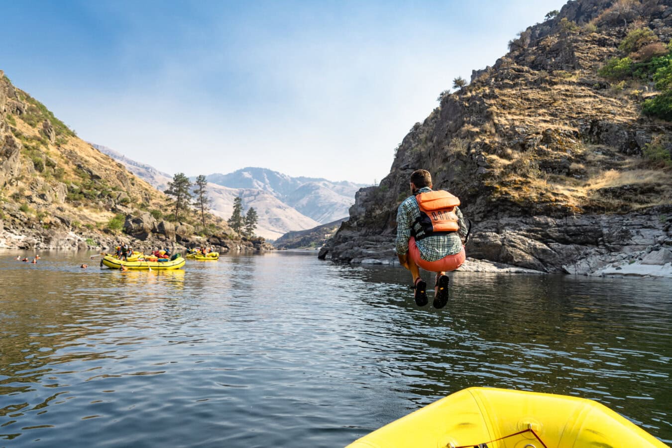 A man wearing an orange life jacket does a cannonball off of a yellow raft into a calm stretch of river