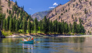 A dory floats down the Salmon River with a tree studded shoreline and mountains in the background.
