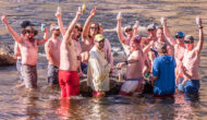 A group of men standing in the Tuolumne River smile and raise their beverages in the air while looking at the camera
