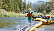 Three inflatable kayaks are turned upside down and lashed together front to back to a large oar raft as an obstacle course game; a young girl is jumping mid-air off of it into the river.