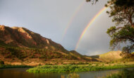 A double rainbow is seen above a riverscape with mountains studded with lots of green vegetation.