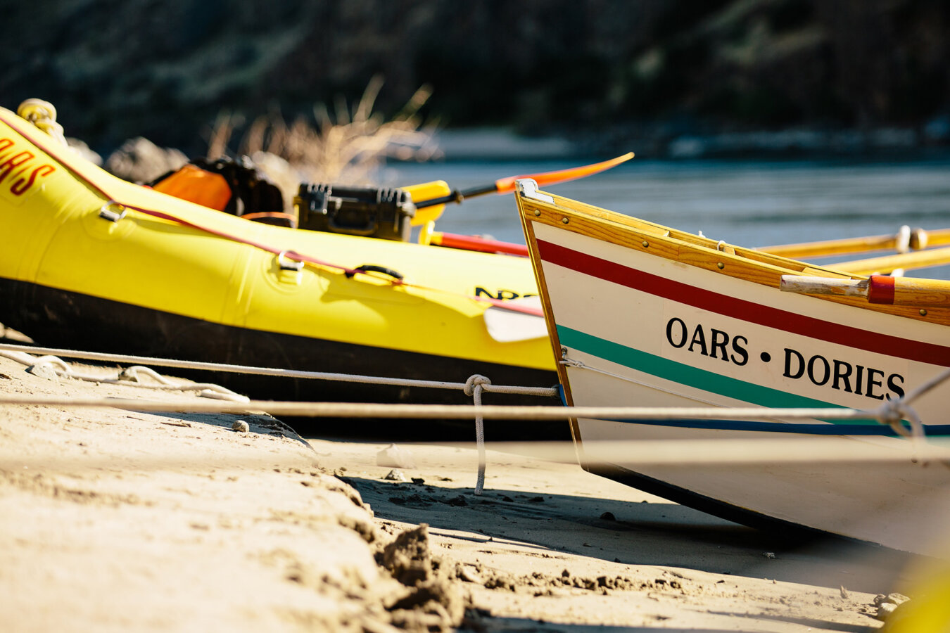 A ground level view close up of a dory and yellow raft tied up on a sandy shore along the river.