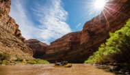 A raft of people floats on a flatwater section of the San Juan River between red canyon walls rising above them.
