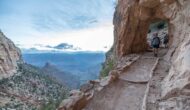 2 people walking through a hole in the rock on a trail next to a cliff.