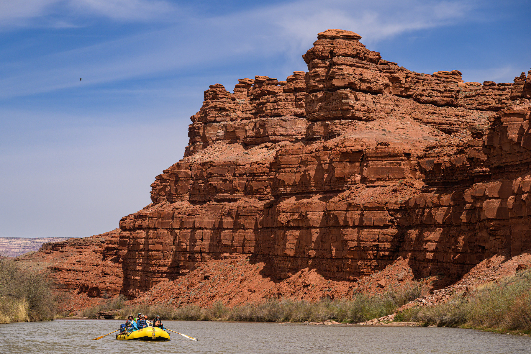 A single yellow raft on the San Juan River floats beneath a towering red rock cliff wall