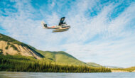 A water plane flies over the river as it takes off surounded by lush, green forested hills.