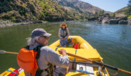 A training guide sits on the front of the raft and watches as a guest rows the raft with oars