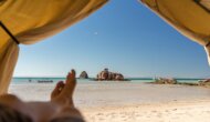 A serene beach view from inside a tent, showcasing turquoise waters, sandy shores, and rock formations under a clear blue sky.