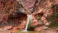 A waterfall spills over a ledge from a vibrant red rock canyon