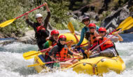 A group of excited rafters in a yellow raft hold their paddles in the air and smile for the camera