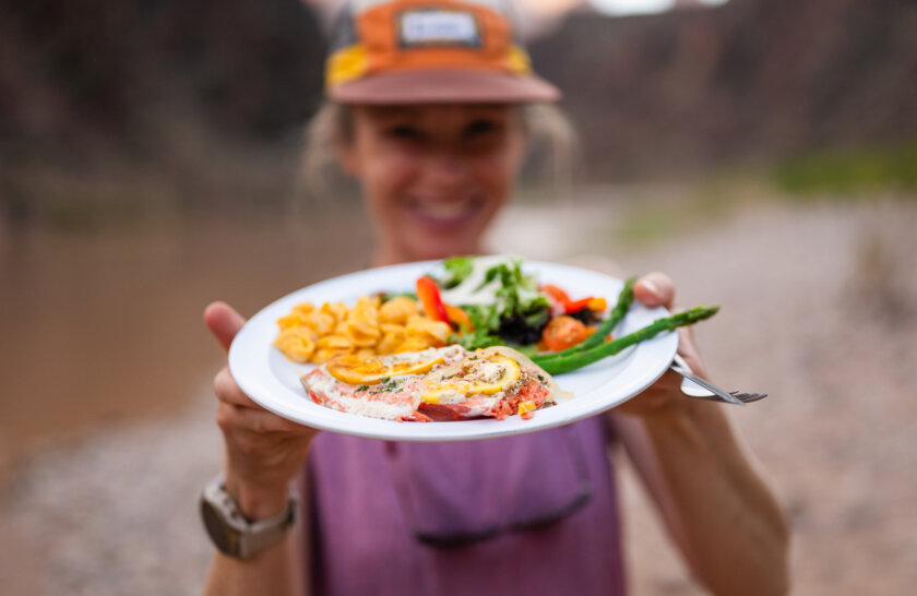 A woman smiles and holds up a plate of salmon, salad, asparagus and mac n cheese.