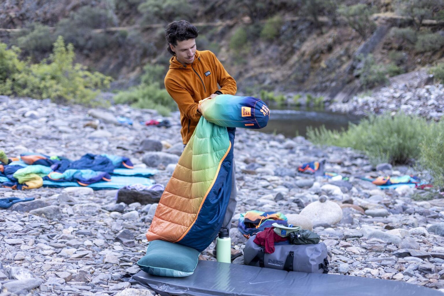 A man stuffs a colorful Rumpl Wrap Sack sleeping bag into a stuff sack on a rocky beach camp along the Tuolumne River.