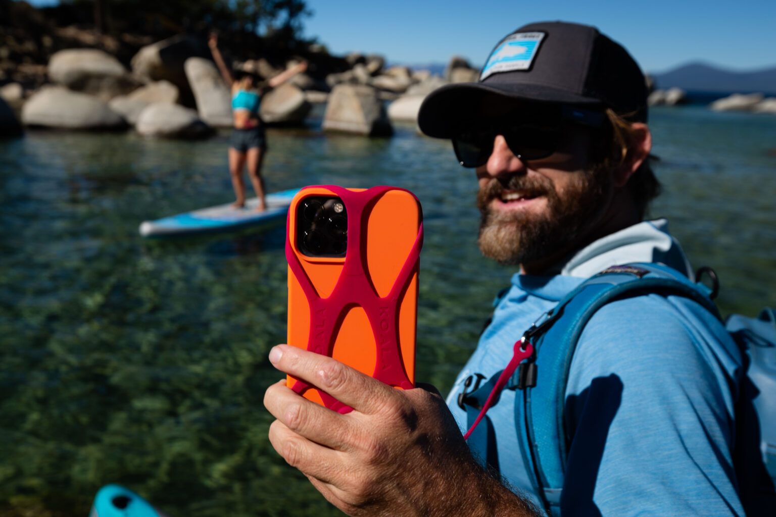 A man holds an iphone that's protected in a special harness to prevent him from dropping the phone as he takes a selfie of himself and his paddle partner on a lake 