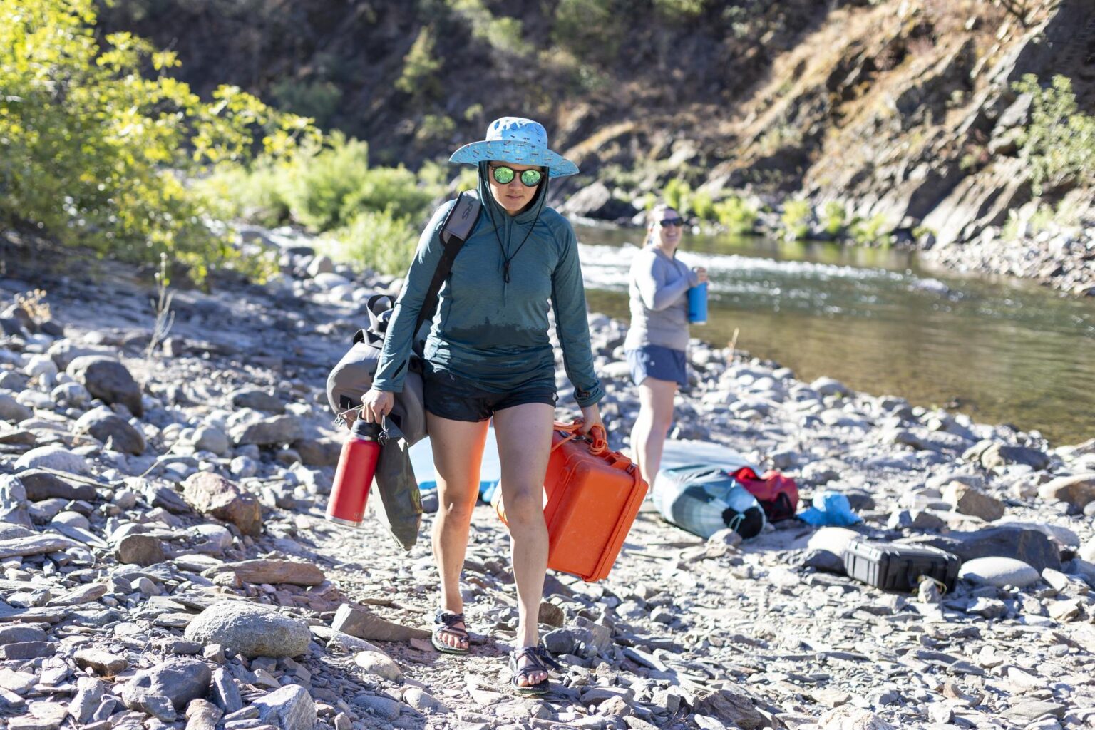 A woman walks along shore with various river gear in our hands