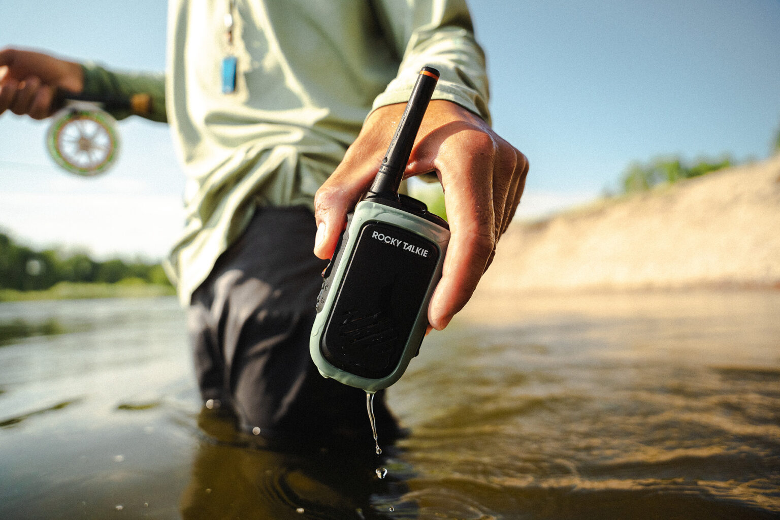A fisherman submerged in water dunks a Rocky Talkie in the water to show how its waterproof