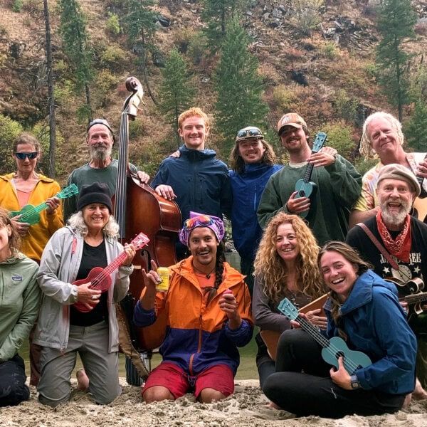 A group of guides and guests pose on a sandy beach along the river and smile for the camera while showing off a variety of instruments on a music river rafting tirp