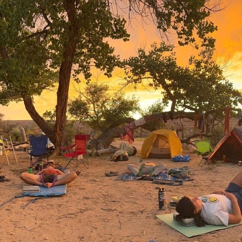 A group of rafters lie on mats on a sandy beach and do yoga with a vibrant sunset over camp.