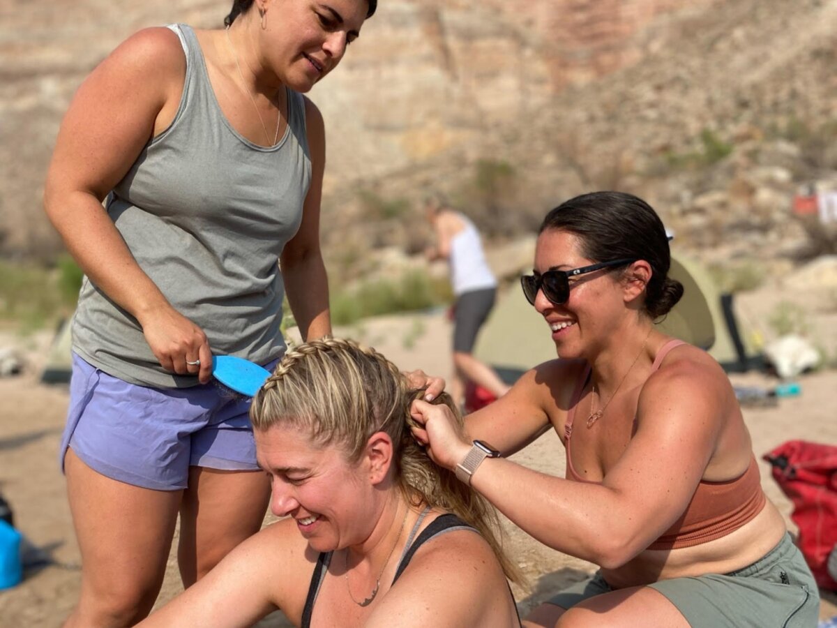 Two women help braid another woman's hair on a sandy beach on the river.