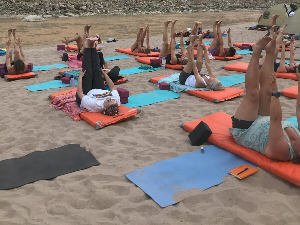A group of women practice yoga on a sandy beach next to the river.