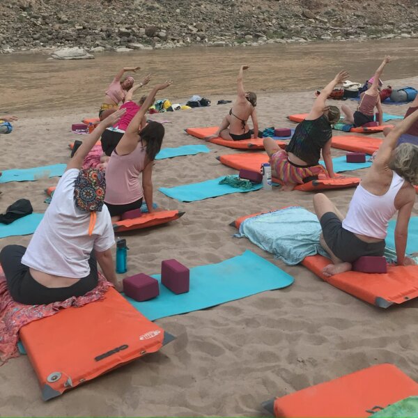 A group of women practice yoga on a sandy beach next to the river.