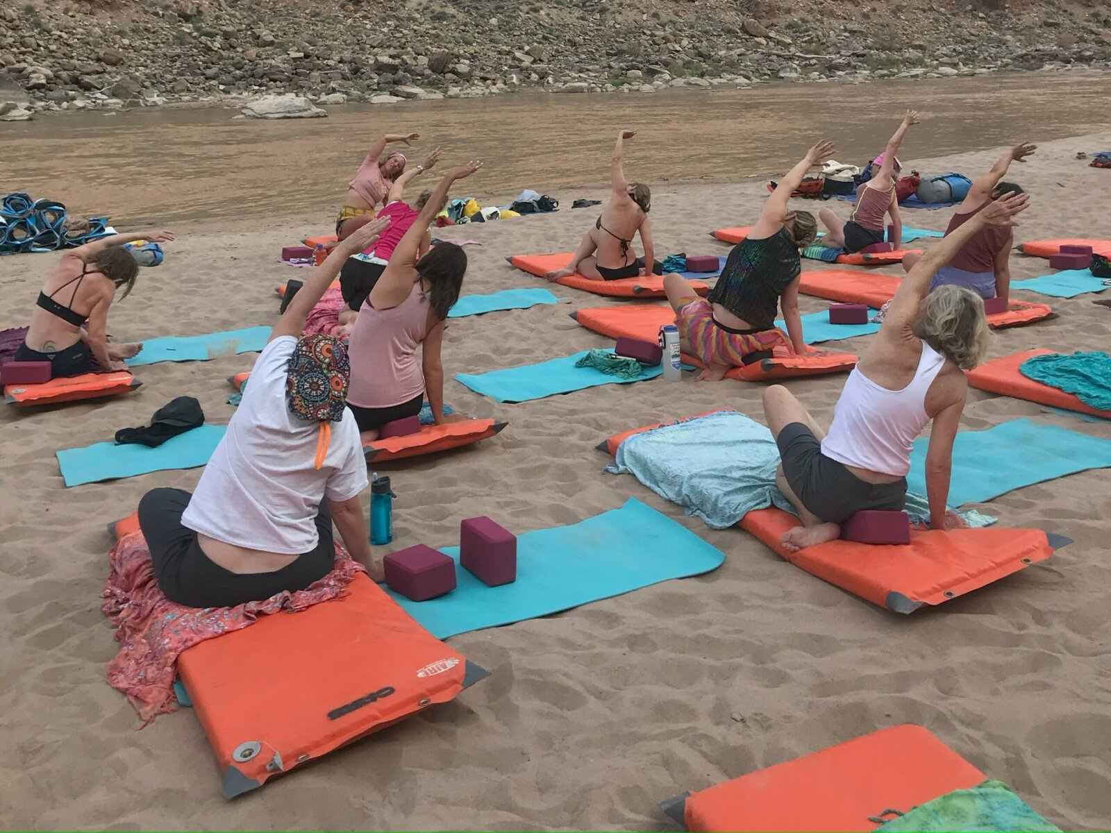 A group of women practice yoga on a sandy beach next to the river.
