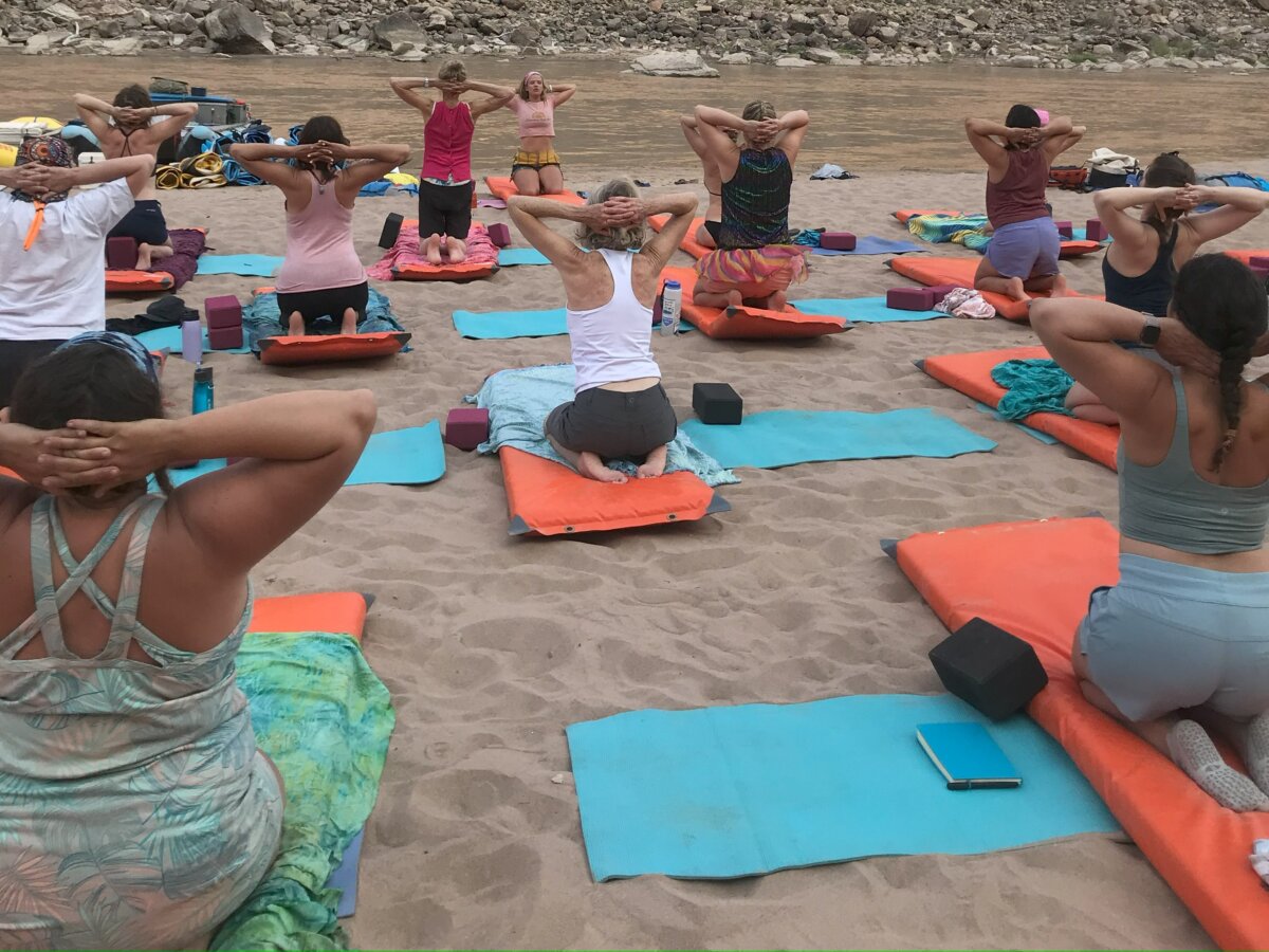 A group of women practice yoga on a sandy beach next to the river.