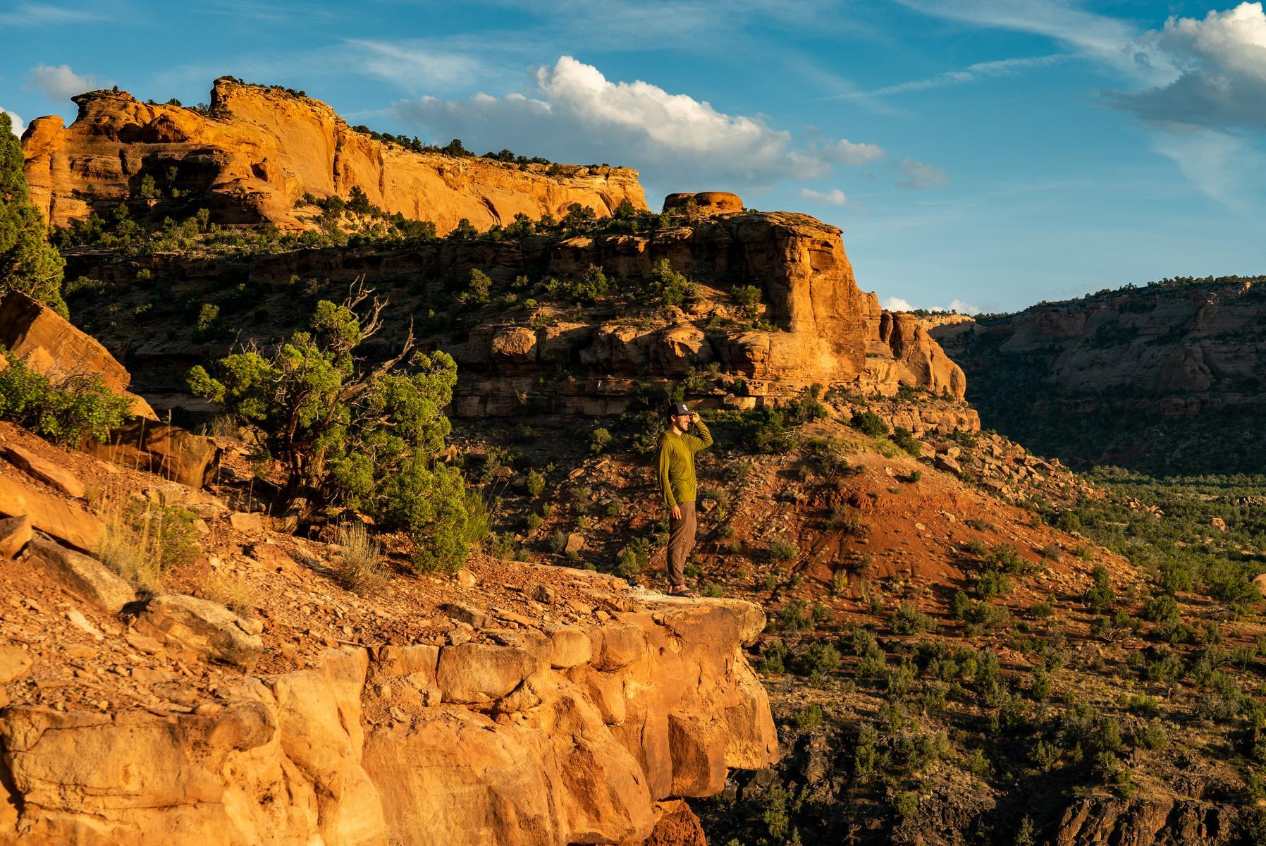 A smiling man stands at the edge of a ledge looking out over a glowing Westwater Canyon as the sun sets.