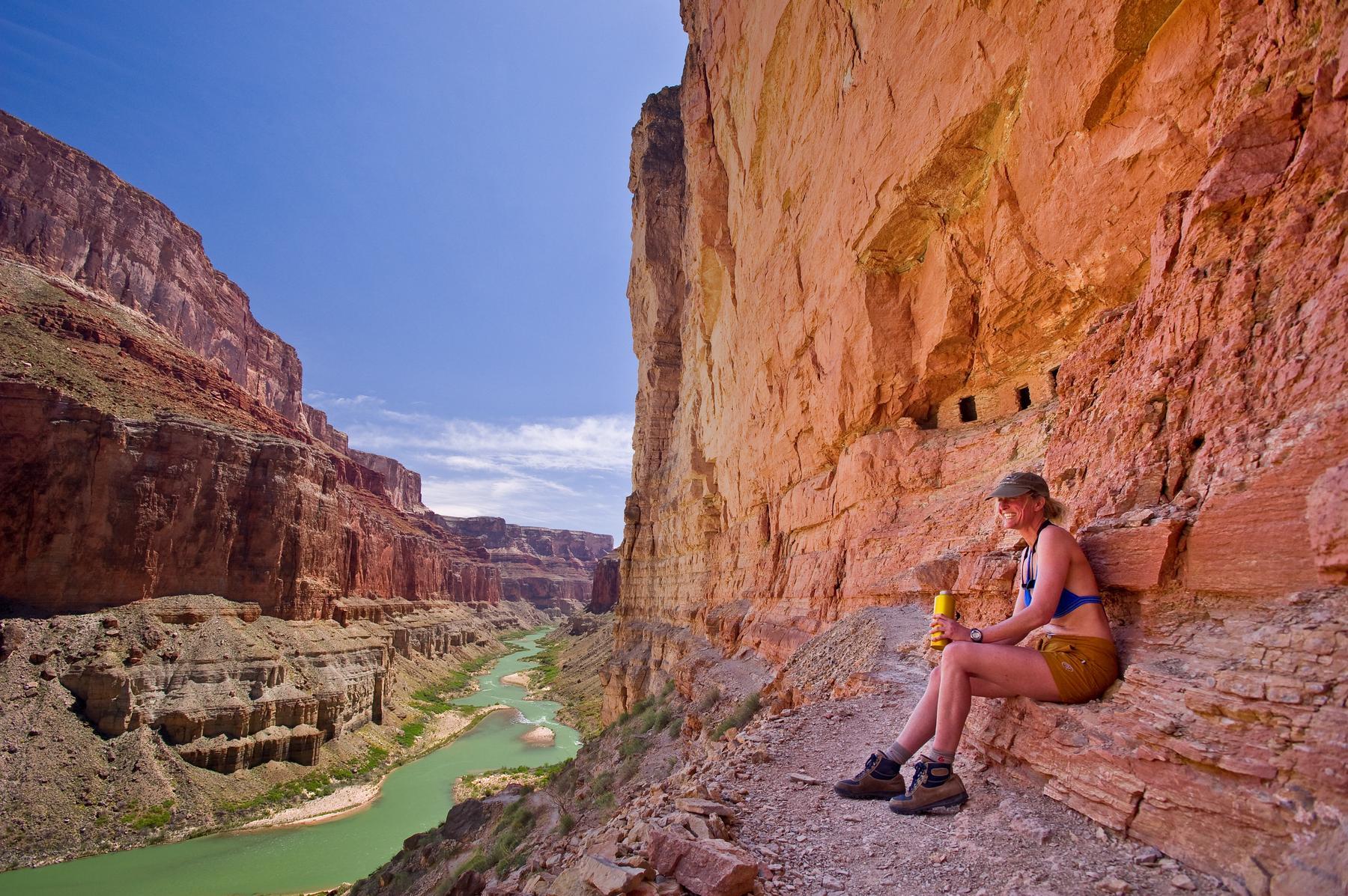 A woman sits in front of the Nankoweap Granaries nestled above the Colorado River in Grand Canyon