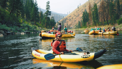 A woman wearing a PFD and helmet sits in an inflatable kayak amongst a group of yellow rafts on the Main Salmon River