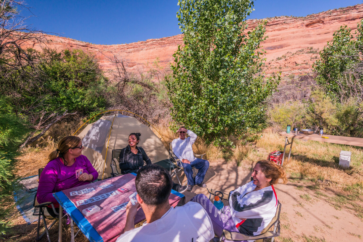 A group of campers smile around a table as they play cards next to a tent at a sandy camp along the Colorado River