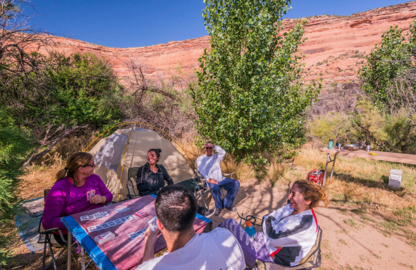 A group of campers smile around a table as they play cards next to a tent at a sandy camp along the Colorado River