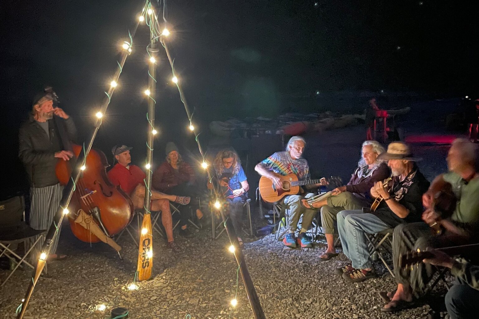 A group of musicians play acoustic instruments on a rocky beach on the river at night while guests watch and three oars are set up with solar string lights.