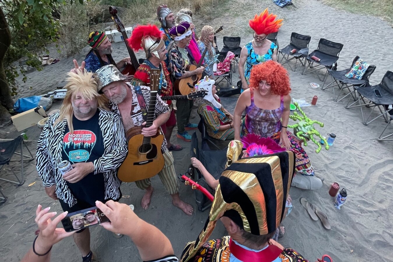 A group of people in vibrant and silly costumes, some holding accoustic instruments, all smile for photos while at camp on an OARS river trip.