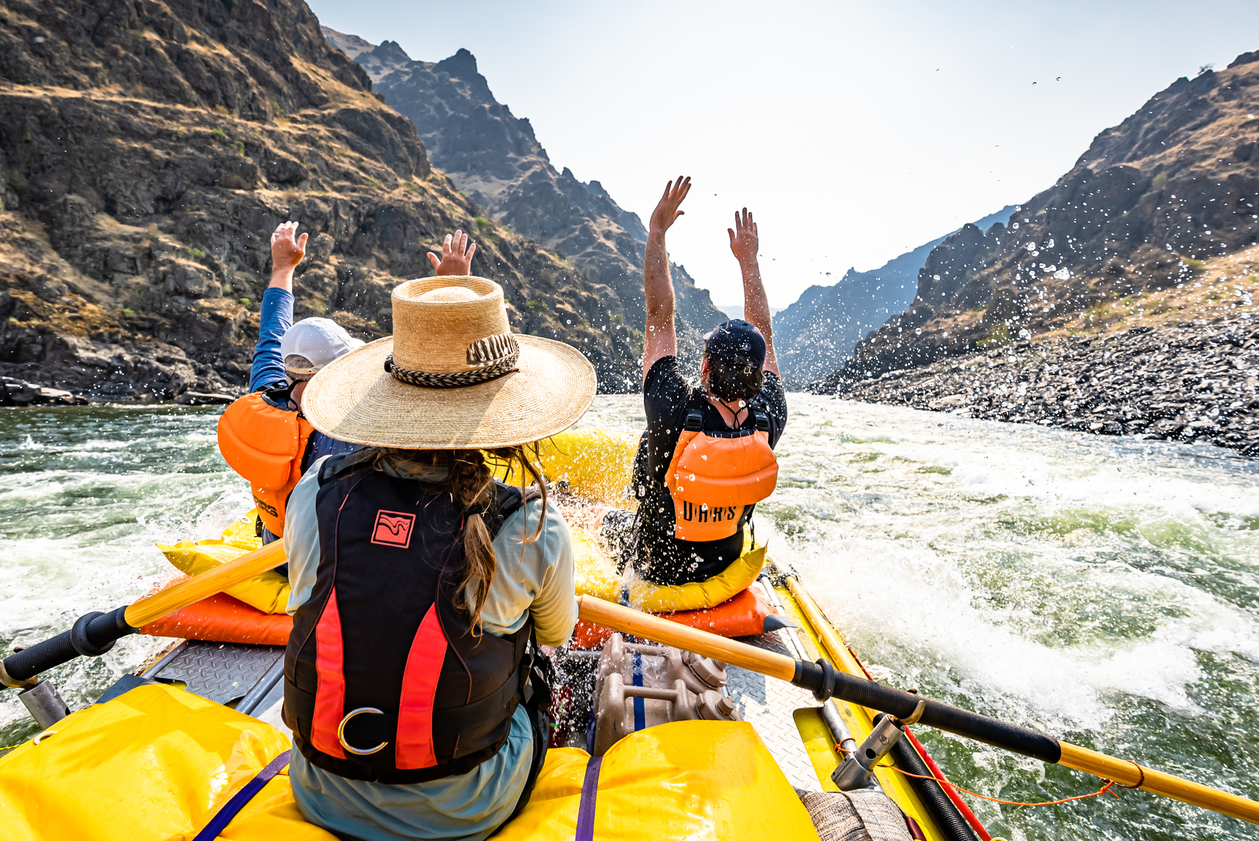 Two people sitting in the front of a yellow raft raise their hands in the air as they are rowed through a rapid by a guide on the Lower Salmon River in Idaho