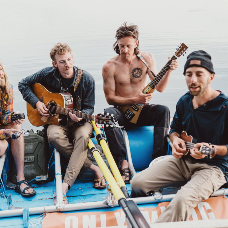 Members of A Brother's Fountain sit on a blue raft on the river and play their instruments.