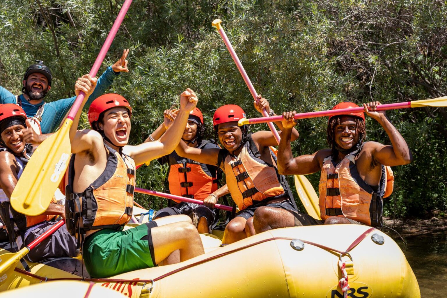 A group of diverse rafters seated in a yellow raft raising there paddles and hands in the air in excitement.